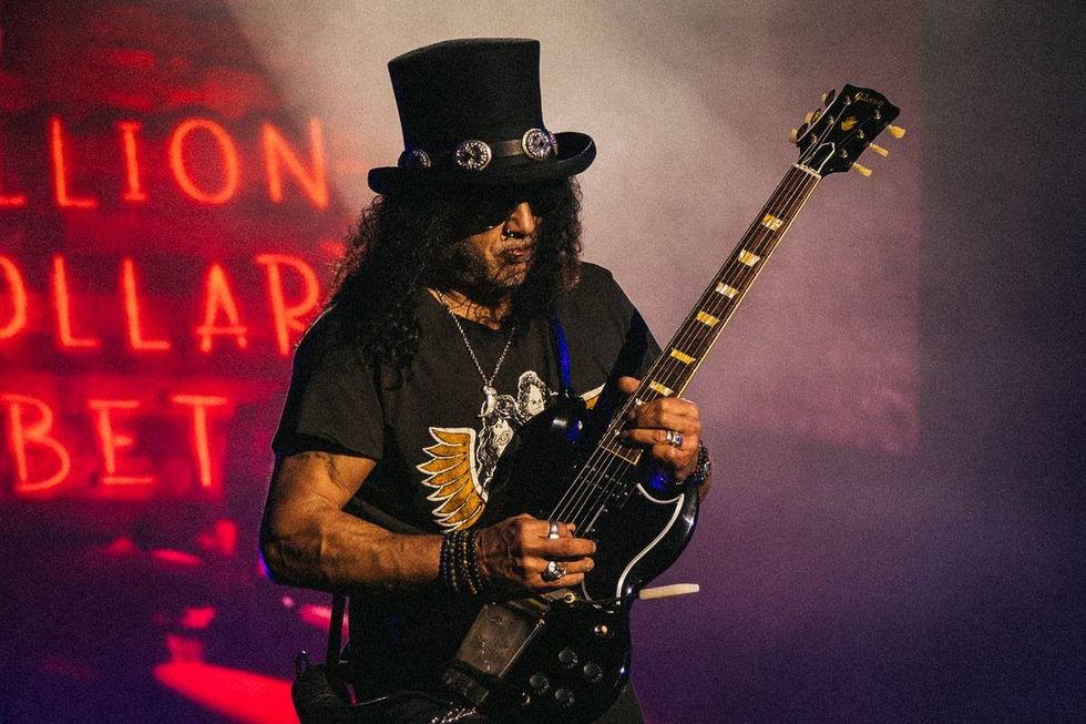 Guitarist in a top hat plays an electric guitar on stage with colorful lighting.