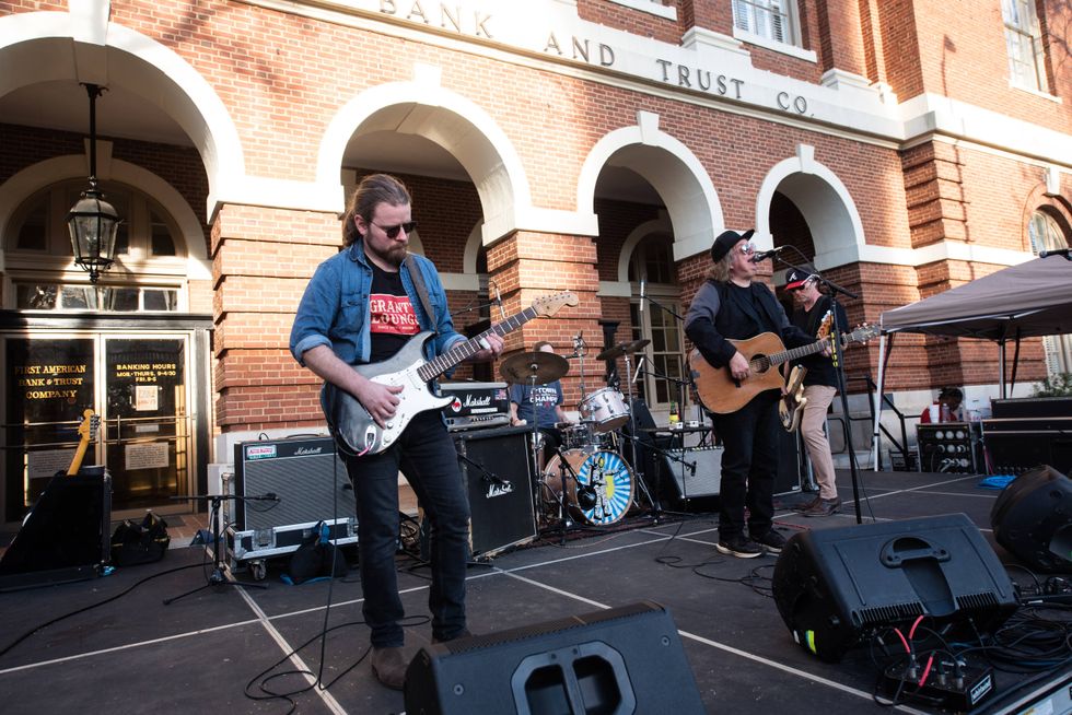 Live band performing on stage in front of a historic bank building, guitars and drums visible.