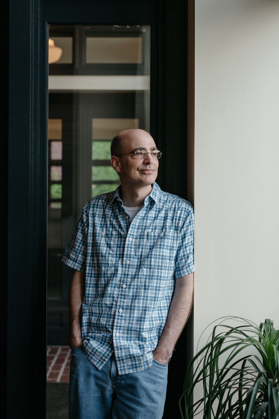 Man in a checkered shirt leans against a doorway, smiling with greenery nearby.