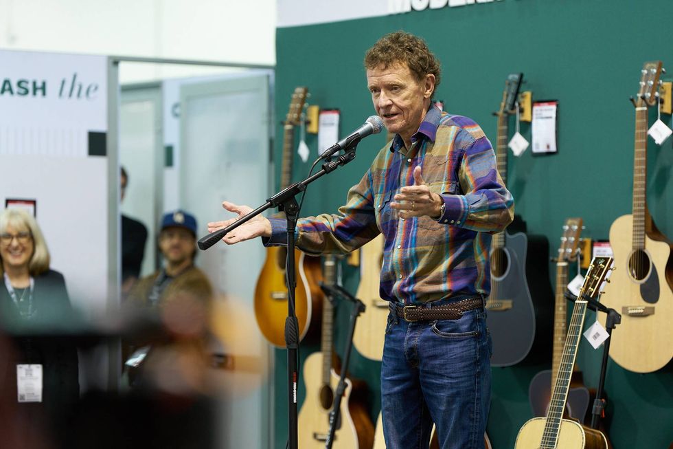 Man in a checkered shirt speaking at a guitar display, engaging an audience.