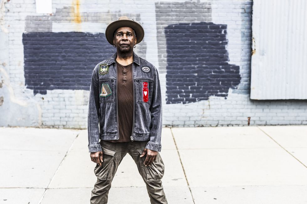 Man in a denim jacket with patches stands confidently against a textured brick wall.