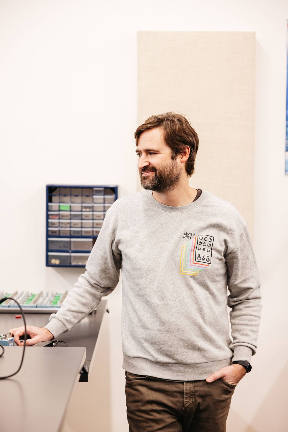 Man in a gray sweatshirt smiles while standing by a workbench with equipment.