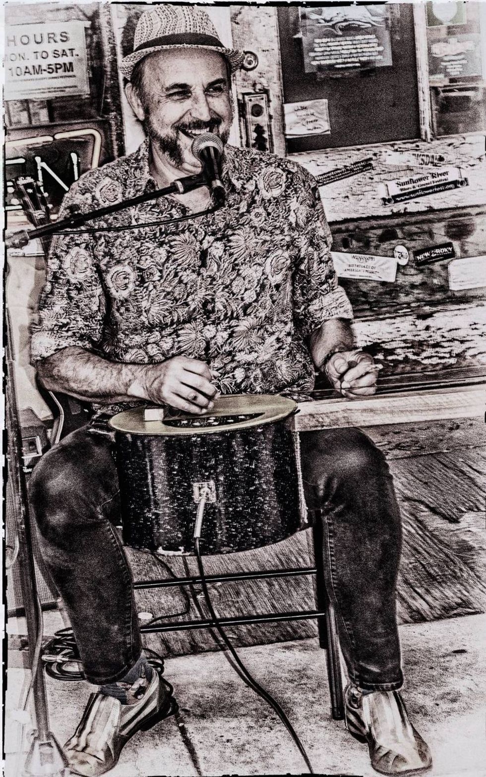 Man in a patterned shirt plays original music, smiling, while seated with a microphone.