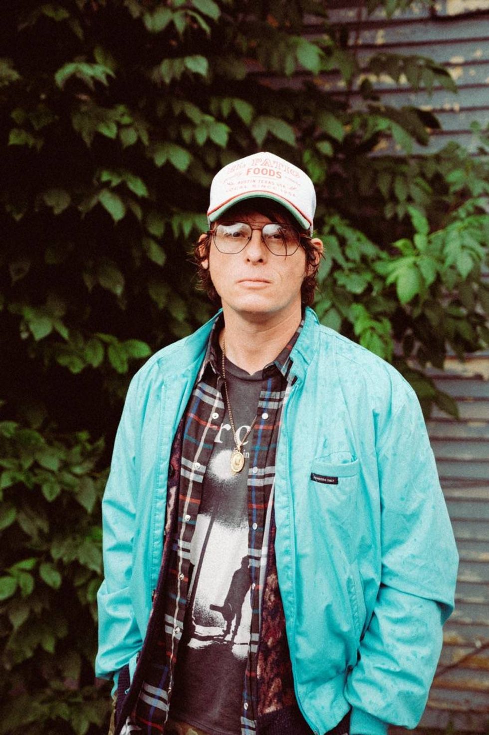 Man in a turquoise jacket and baseball cap stands against a green backdrop.