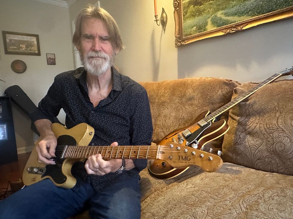 Man playing an electric guitar while seated on a couch, with another guitar nearby.