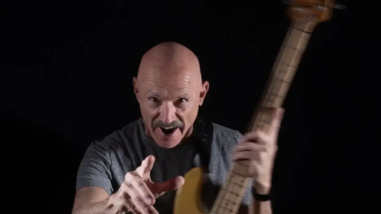 Man with a bald head and mustache passionately playing a bass guitar against a black background.