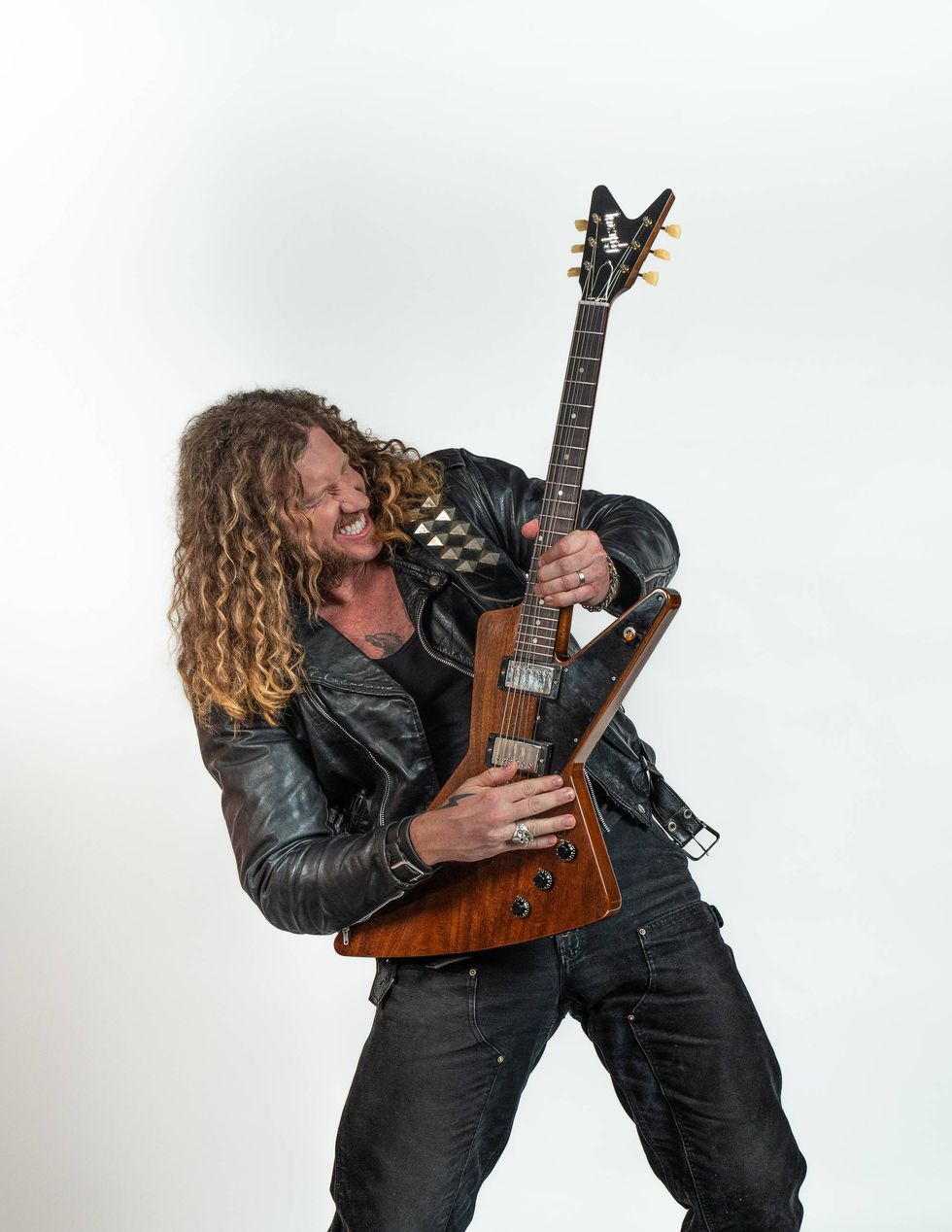 Man with curly hair and leather jacket passionately playing an electric guitar.