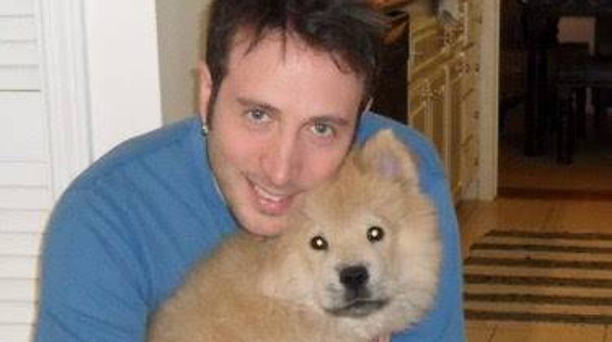 Man with dark hair smiles while holding a fluffy puppy in a home setting.