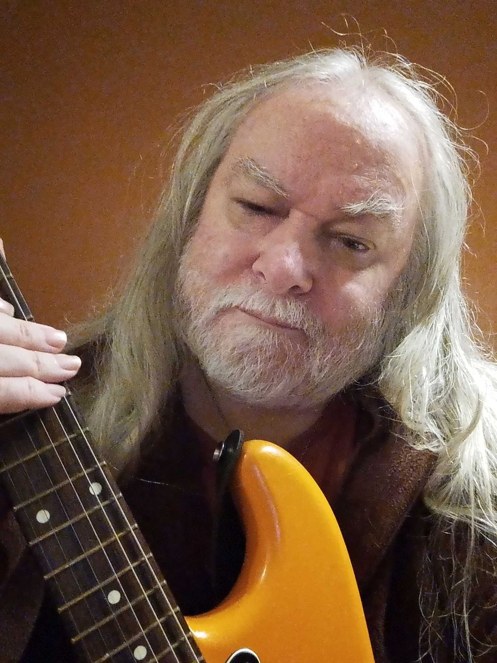 Man with long gray hair and beard, holding an orange electric guitar, gives a playful look.