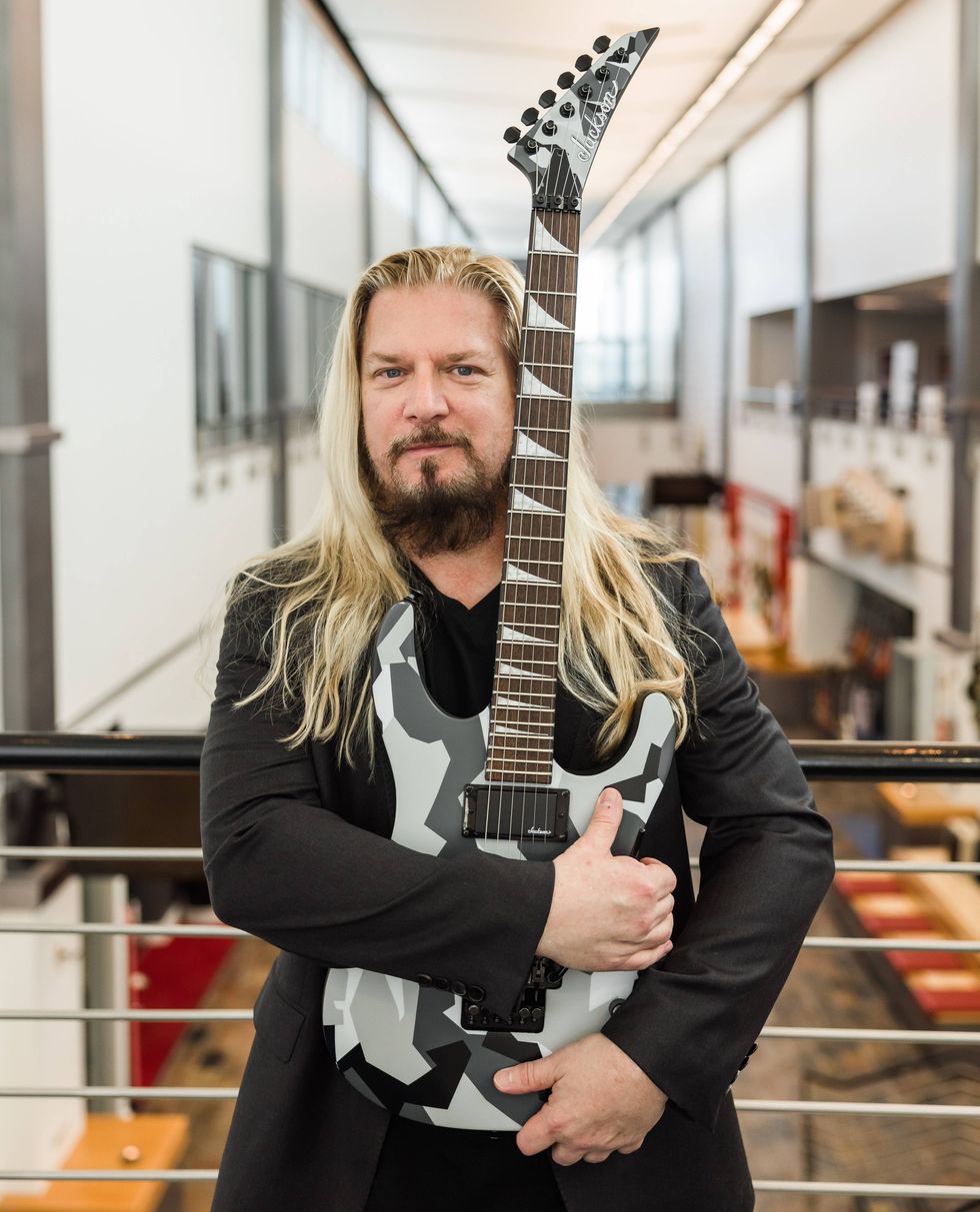 Man with long hair holding a camo-pattern electric guitar in a modern indoor space.