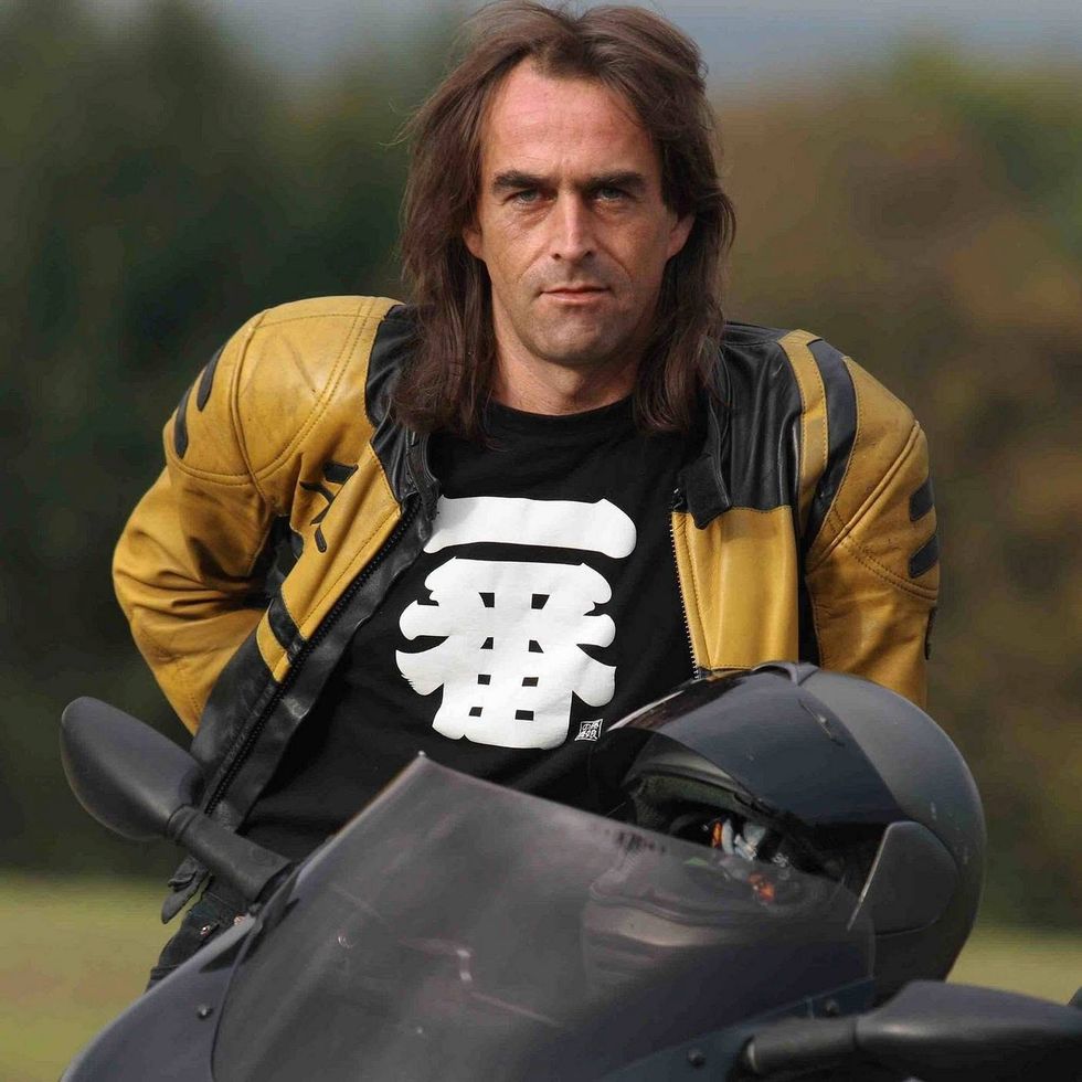 Man with long hair in a yellow and black leather jacket, leaning on a motorcycle.