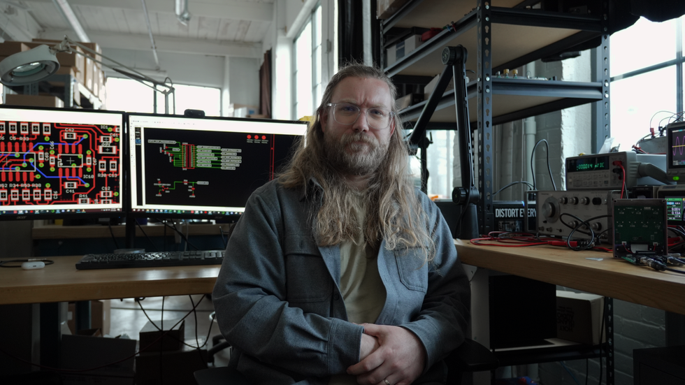 Man with long hair sits at a workstation, surrounded by electronics and dual monitors displaying schematics.