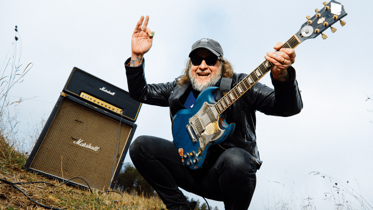 Man with sunglasses and a guitar poses joyfully next to a Marshall amplifier outdoors.