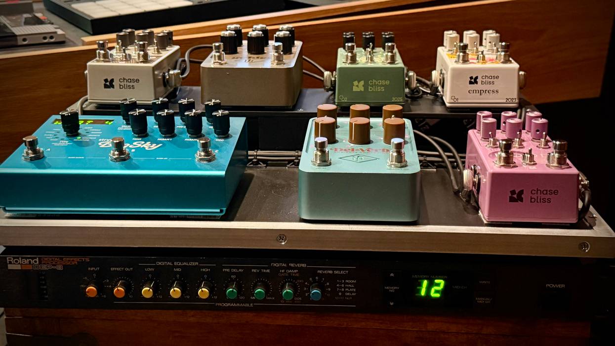 Multiple guitar pedals arranged on a rack in a studio with musical equipment in the background.