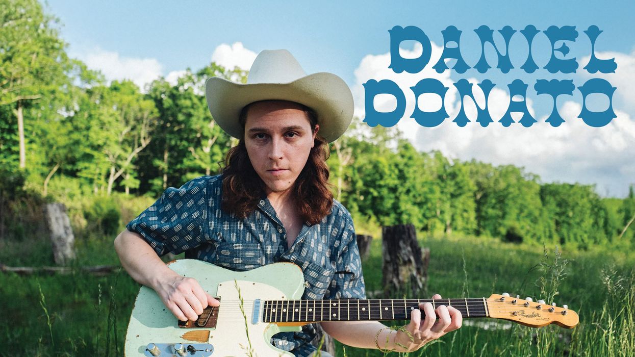 Musician Daniel Donato sits outdoors, wearing a cowboy hat, holding a guitar amid greenery.