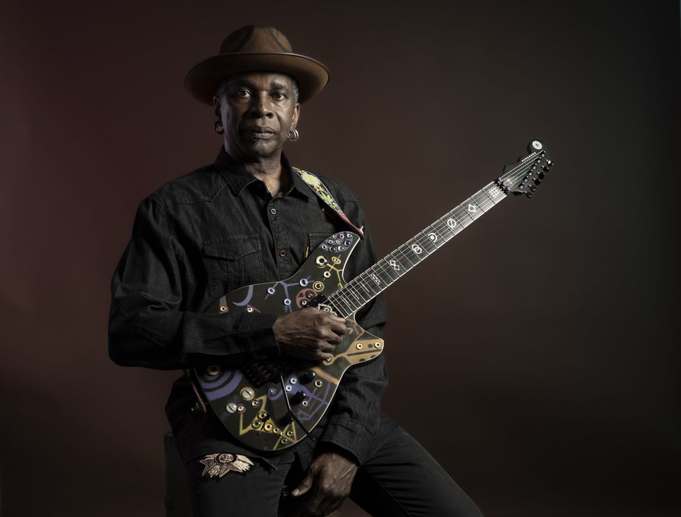 Musician holding a uniquely painted electric guitar, wearing a hat and black shirt.