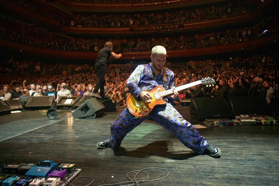 Musician in a floral suit plays guitar on stage; excited crowd in the background.