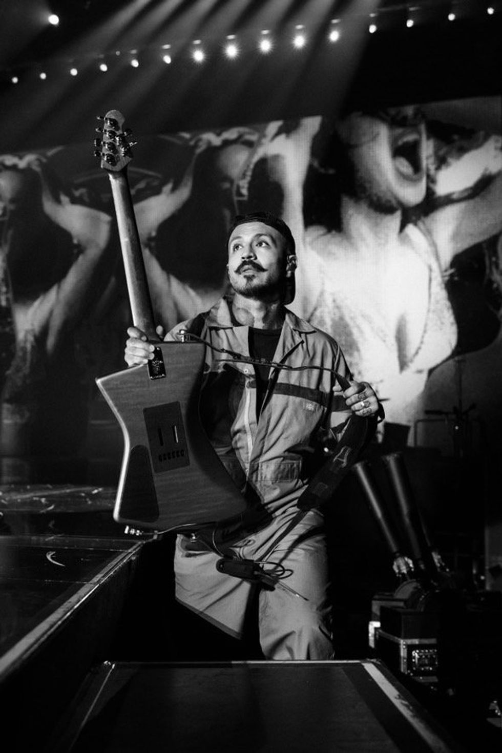 Musician in a jumpsuit backstage, holding a guitar, with dramatic lights and projections behind.