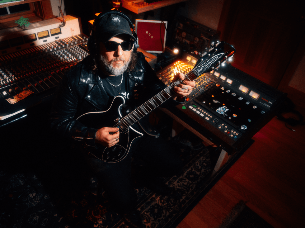 Musician in a leather jacket playing guitar in a dimly lit studio with sound equipment.