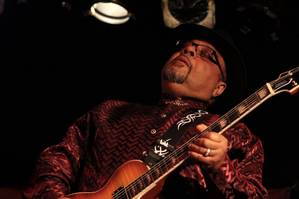 Musician in a patterned shirt plays guitar, eyes closed, lost in the music.