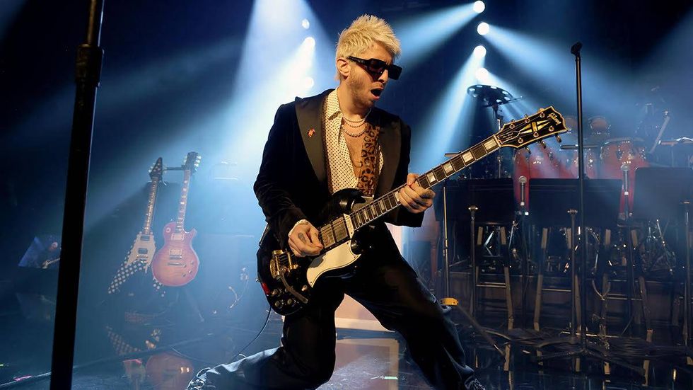 Musician in sunglasses plays electric guitar on stage with dramatic lighting.