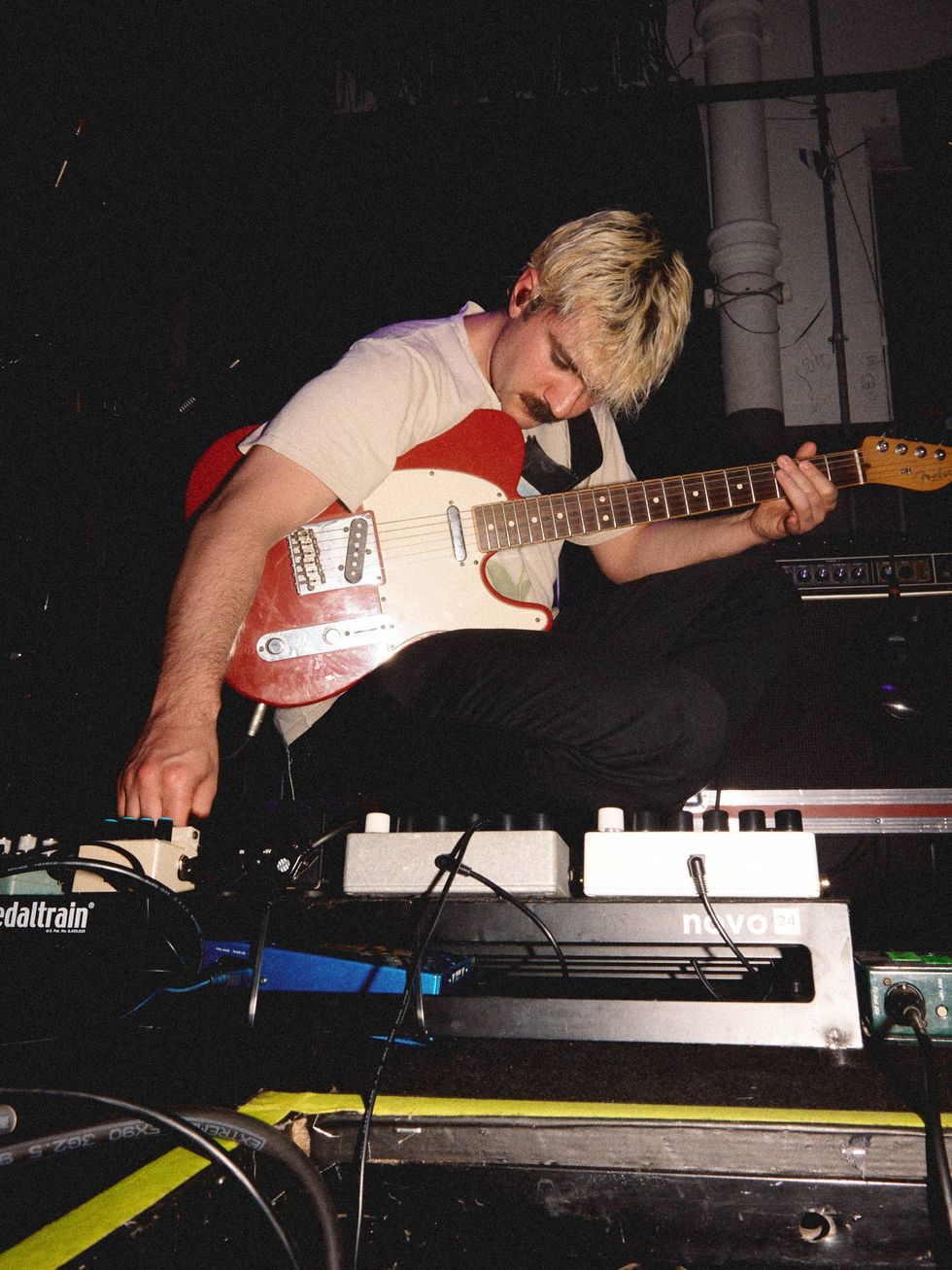Musician kneels on stage with a red guitar, adjusting effects pedals amidst dim lighting.