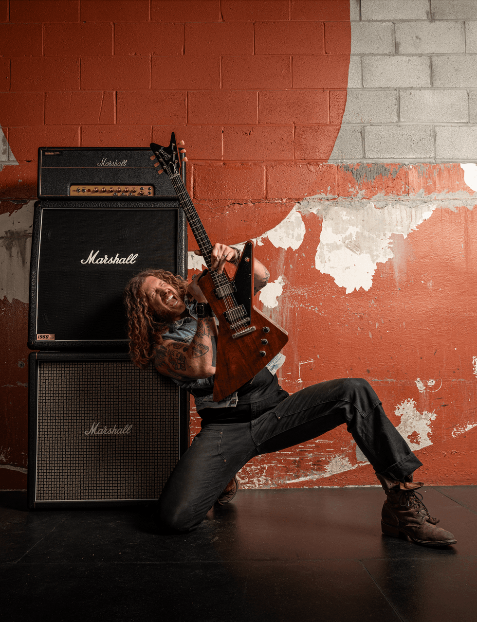 Musician passionately playing a guitar beside towering Marshall amplifiers against a textured wall.
