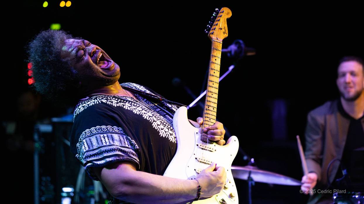 Musician passionately plays guitar on stage, accompanied by a smiling drummer.