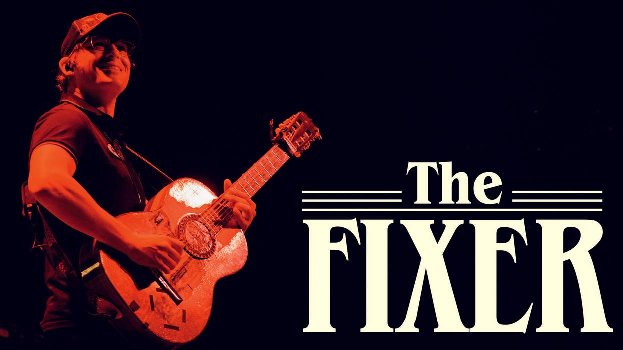 Musician performing on stage, holding a guitar with the title "The Fixer" displayed.