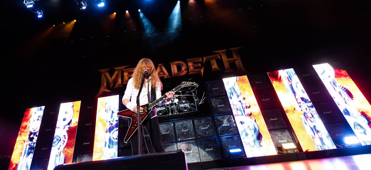 Musician performs on stage with guitar; vibrant lights and band logo backdrop.