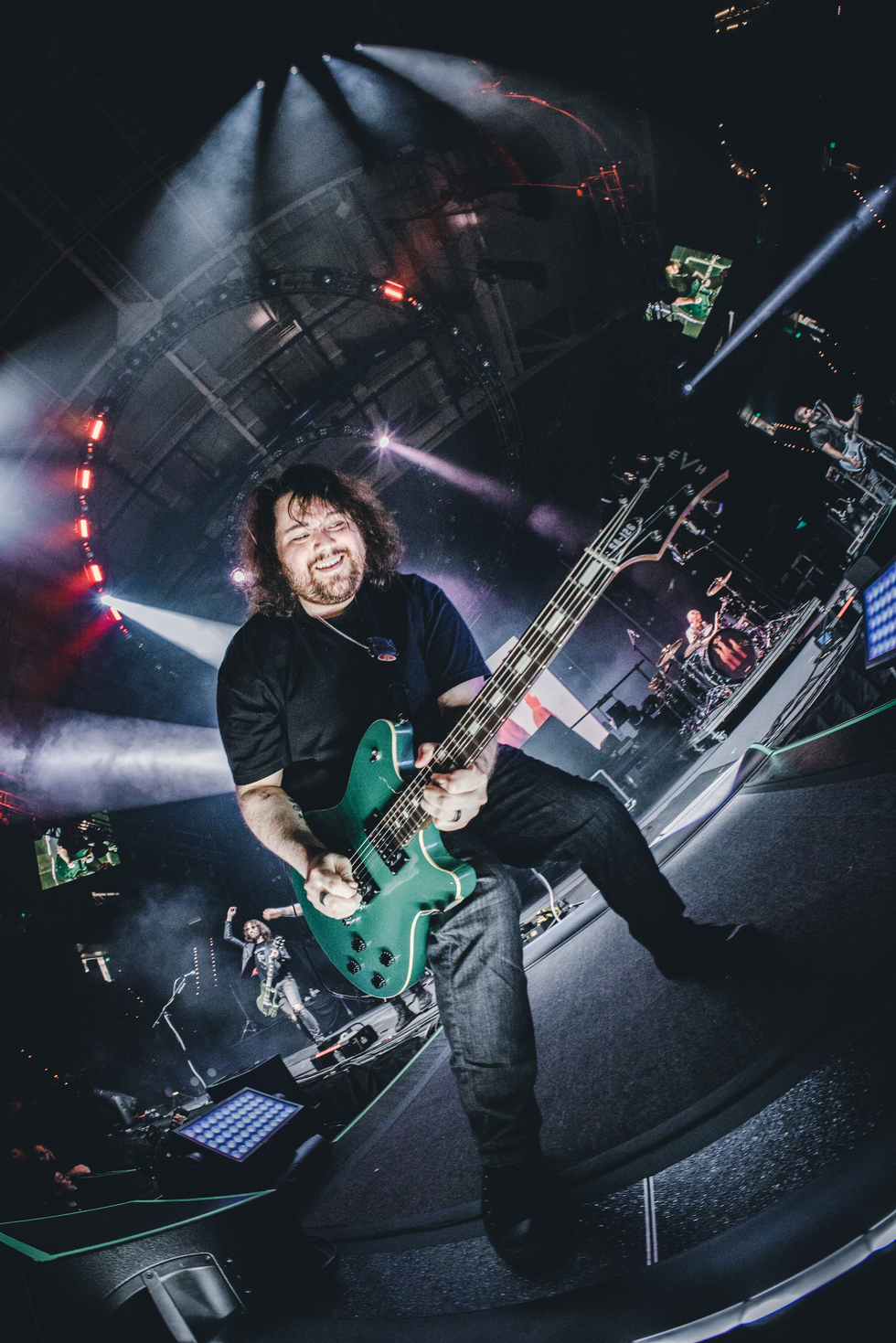 Musician playing a green guitar on stage, smiling, with colorful lights in the background.