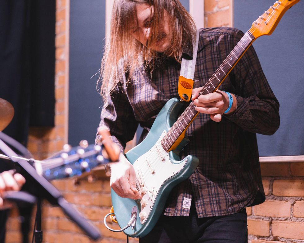 Musician playing a teal electric guitar in a studio setting with brick walls.