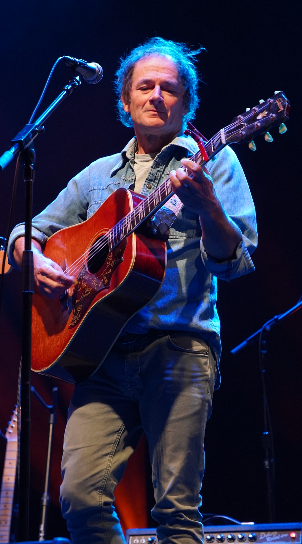Musician playing an acoustic guitar on stage with a microphone and colorful lighting.