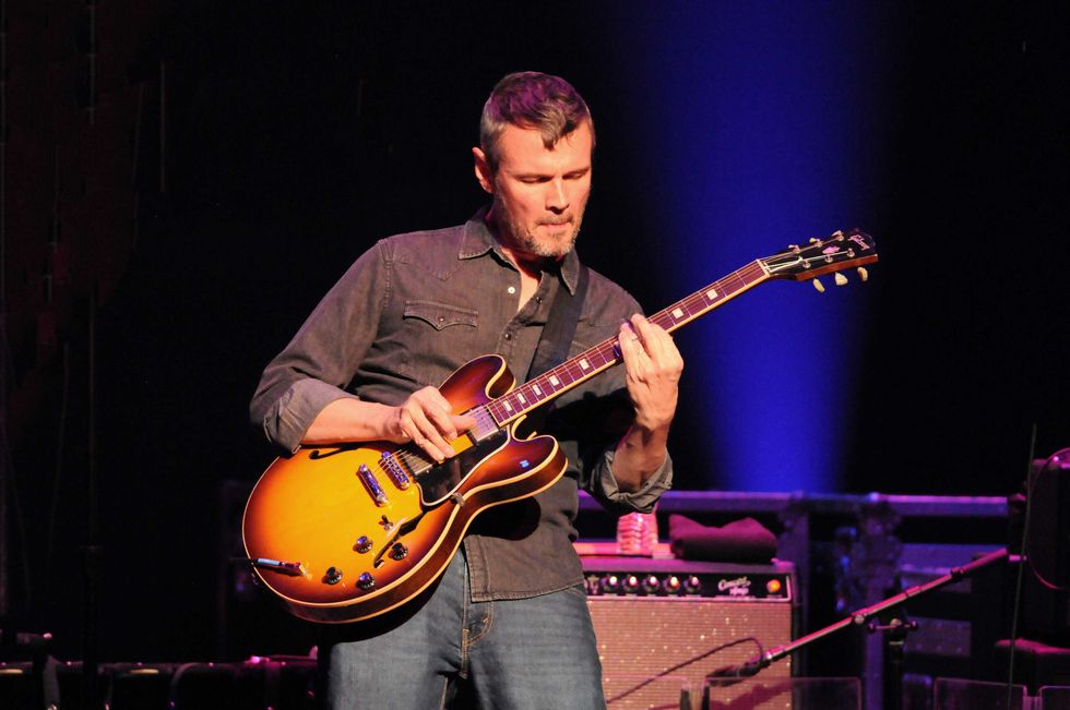 Musician playing an electric guitar on stage, illuminated by colorful lights.