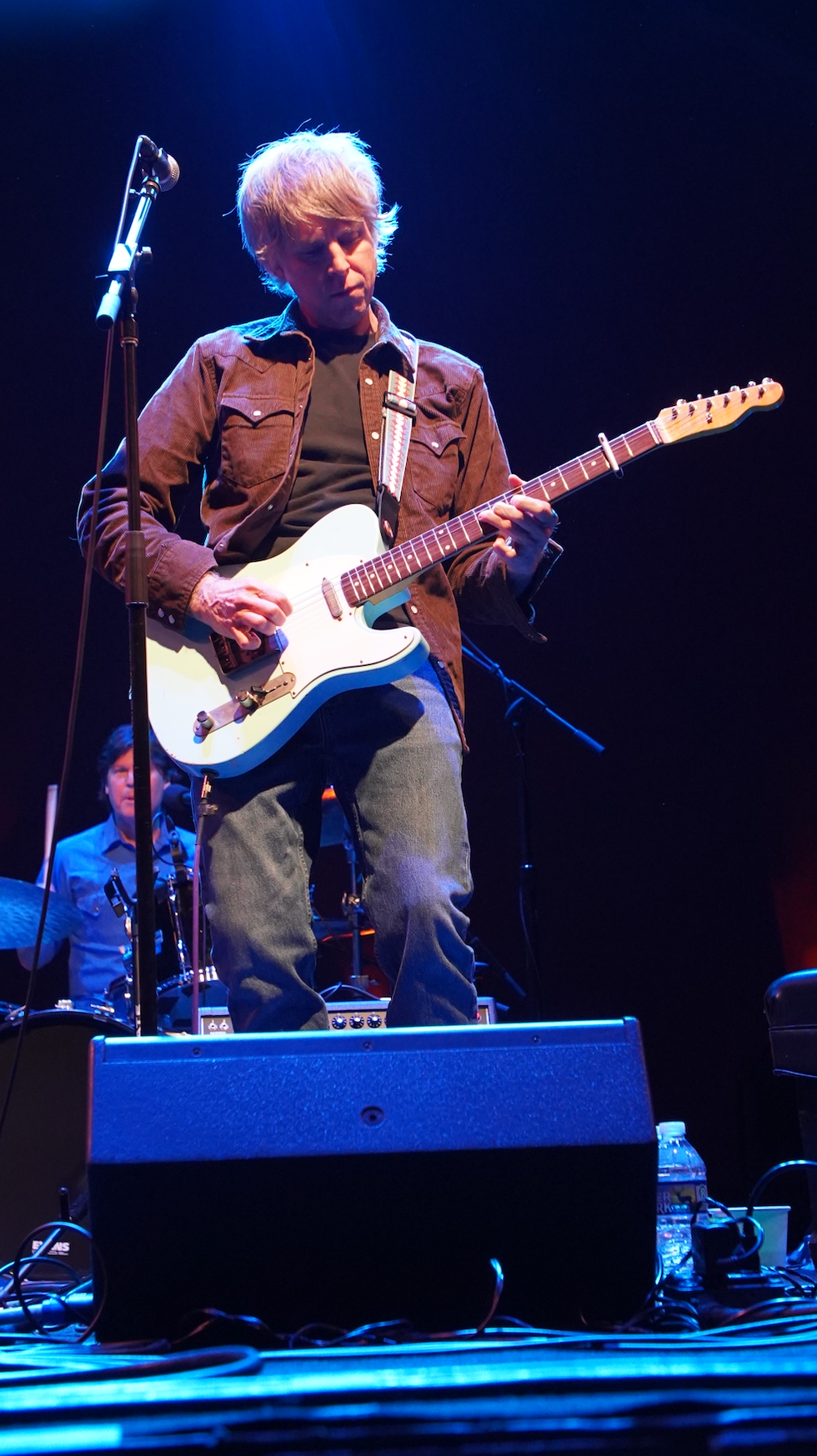 Musician playing an electric guitar on stage with drummer in the background.
