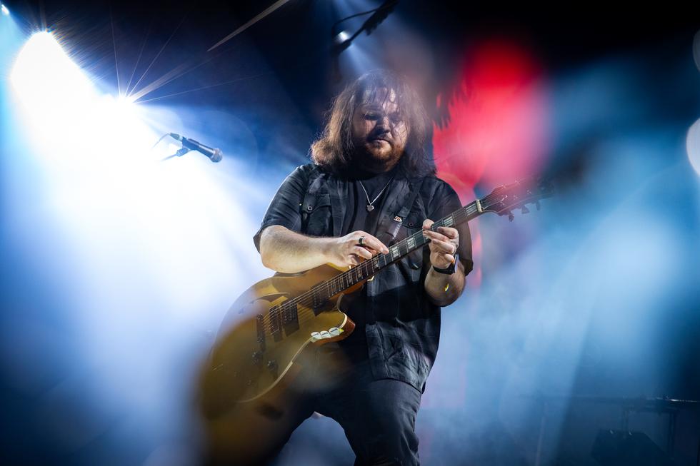 Musician playing electric guitar on stage, illuminated by bright lights and surrounded by fog.