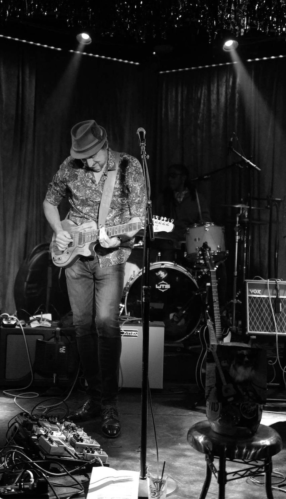 Musician playing electric guitar on stage, surrounded by instruments and equipment.