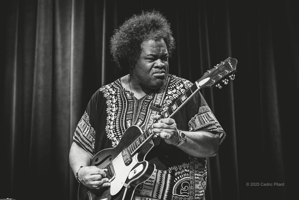 Musician playing guitar passionately on stage, wearing a patterned shirt, in black and white.
