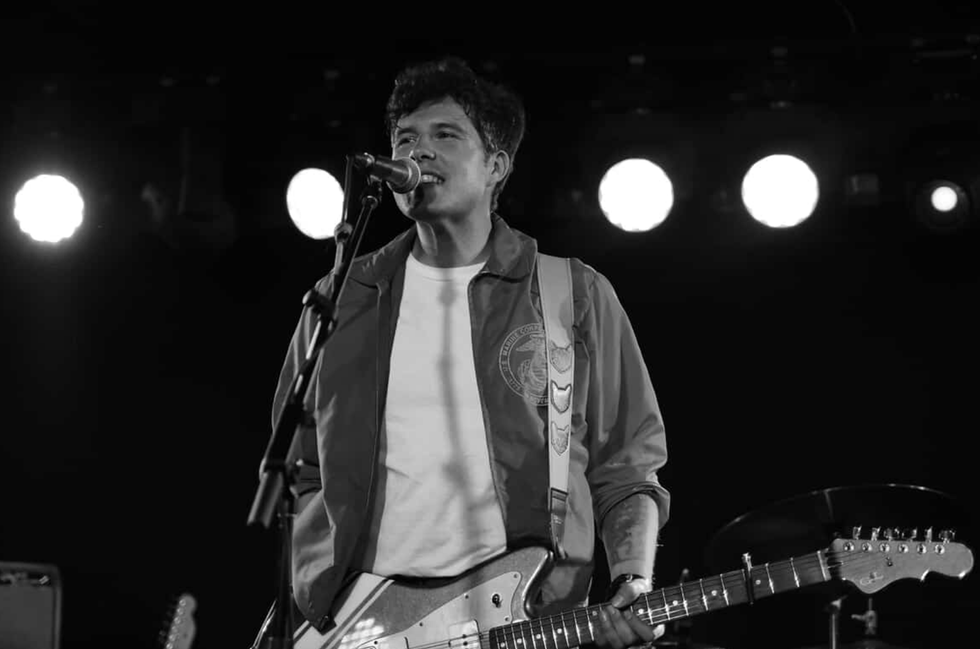 Musician singing into a microphone while playing an electric guitar on stage.