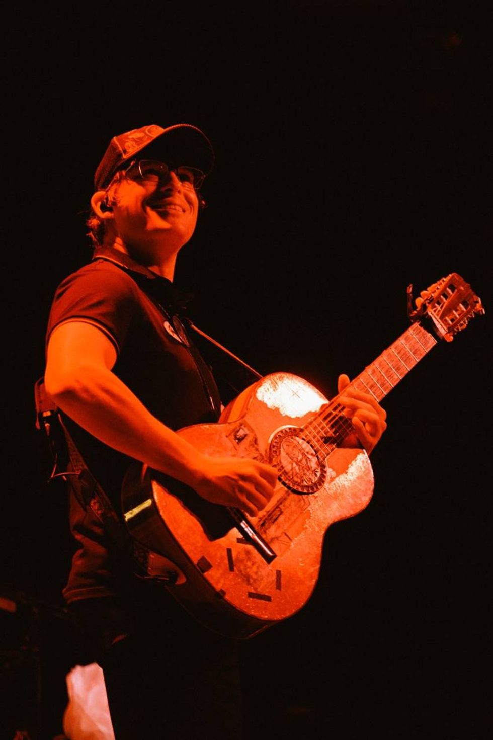 Musician smiling while playing a decorated guitar under red stage lighting.