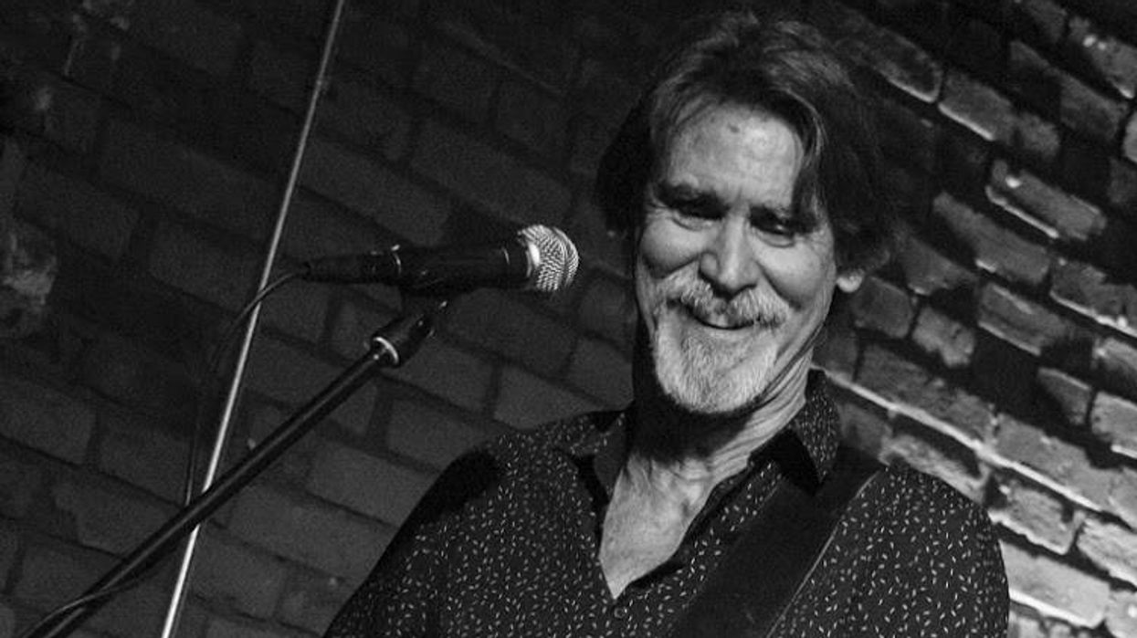 Musician smiling while playing an electric guitar on stage against a brick wall.