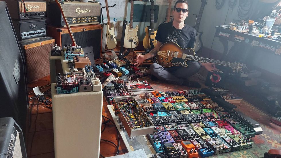 Musician surrounded by an array of guitar pedals and amplifiers, playing an electric guitar.