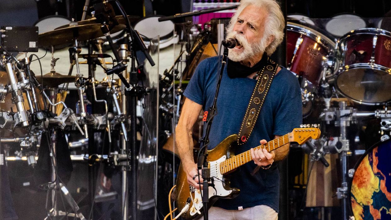 Musician with a guitar singing on stage, surrounded by drums and instruments.