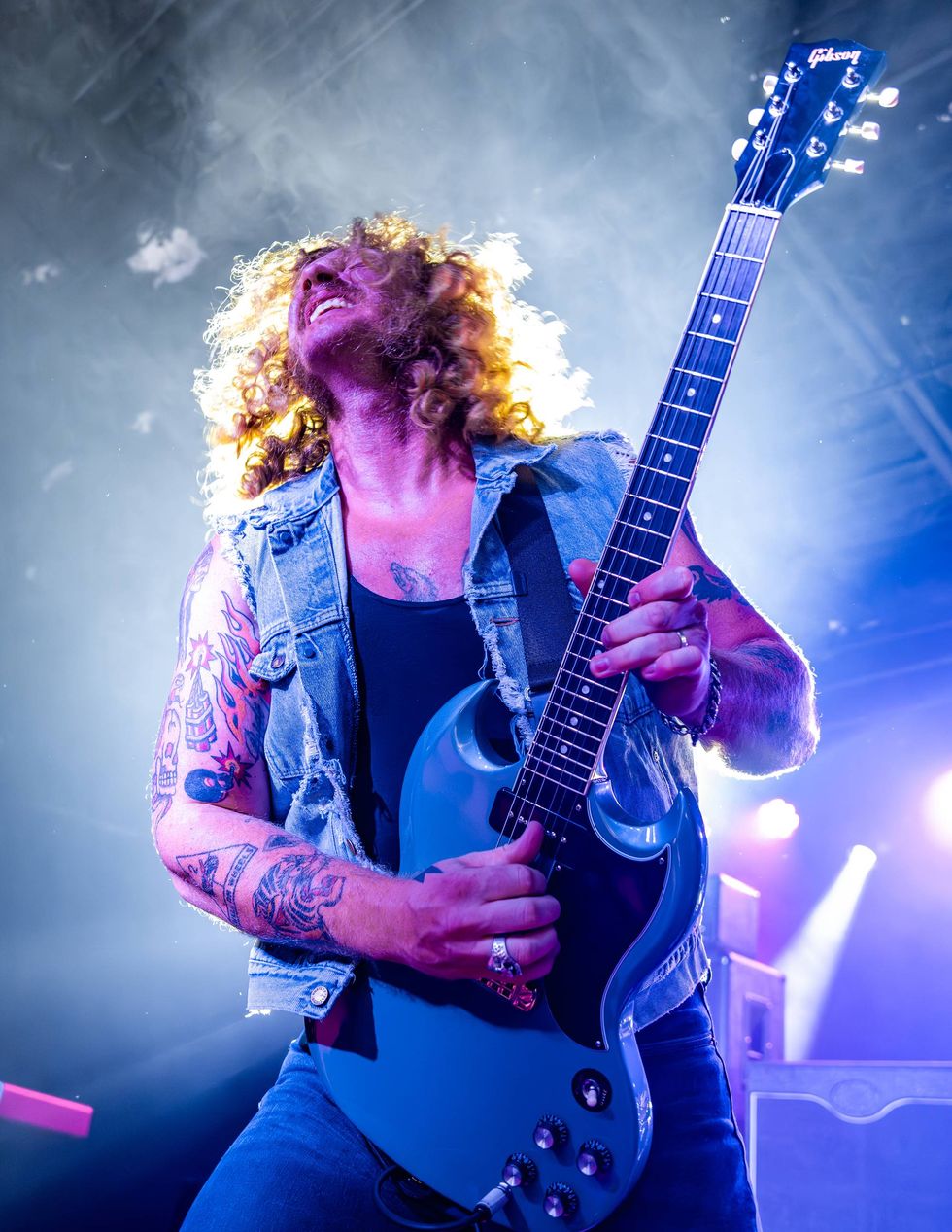 Musician with curly hair passionately plays a blue guitar on stage under colorful lights.