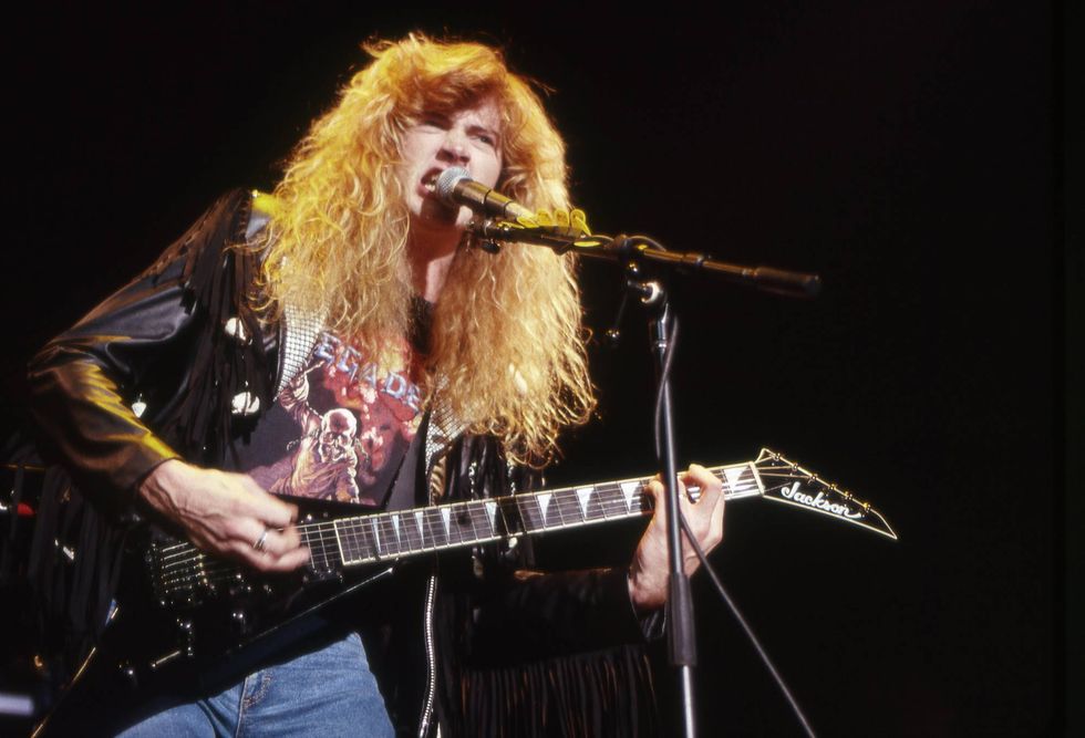 Musician with long hair plays electric guitar on stage, wearing a rock band t-shirt.
