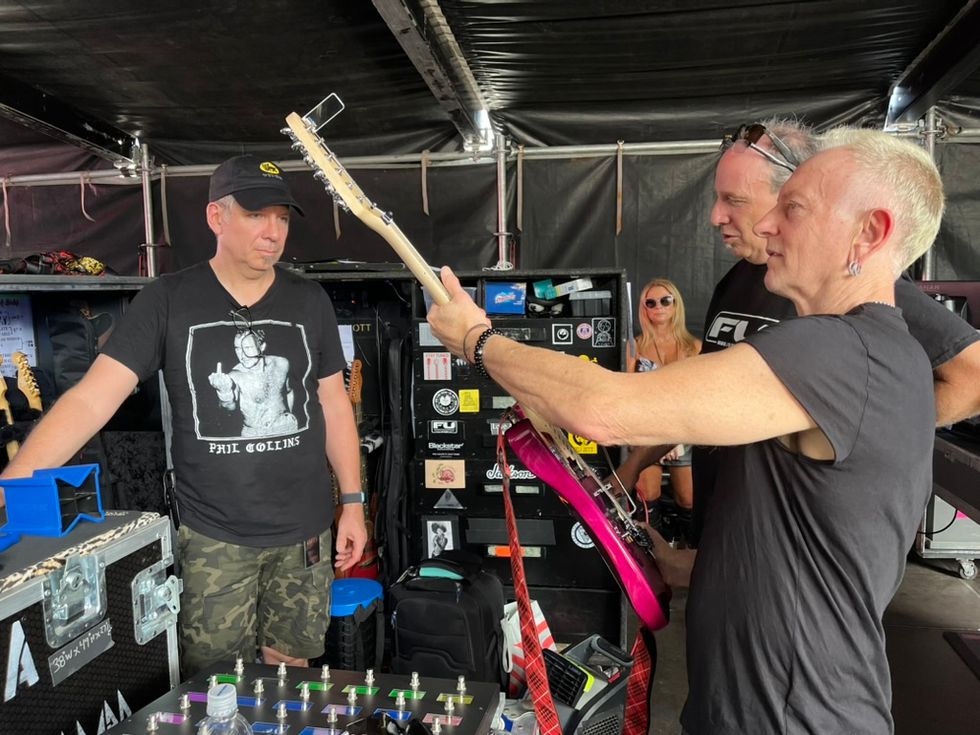 Musicians discussing gear backstage; one holds a pink guitar, others observe.