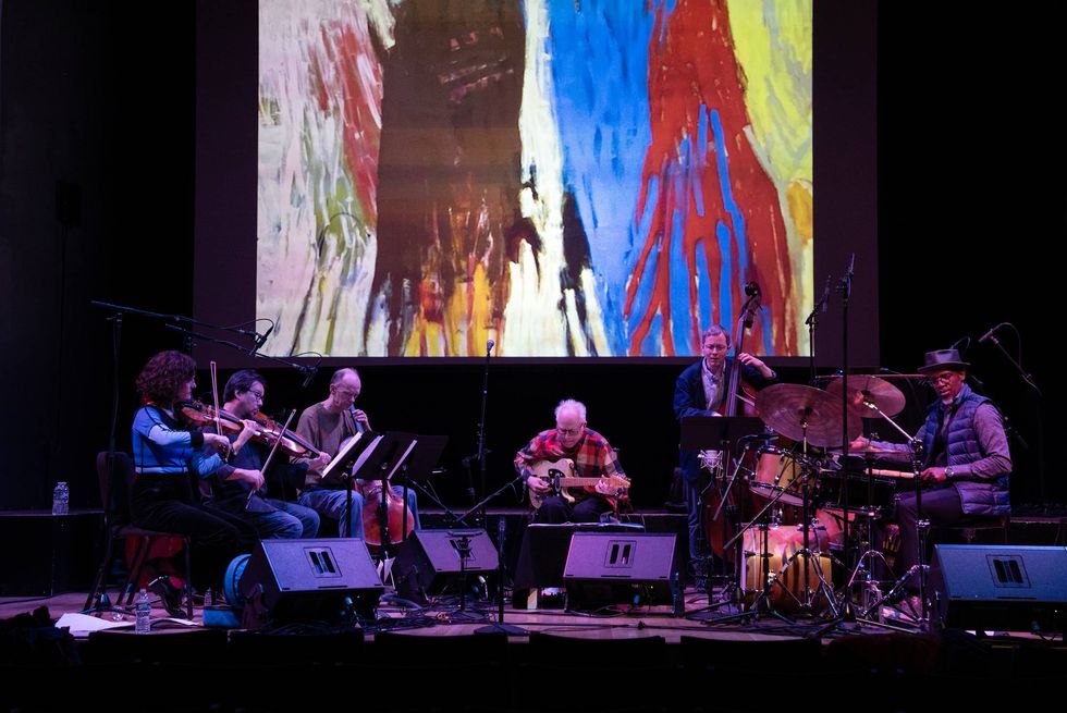 Musicians performing on stage, with colorful abstract art projected behind them.