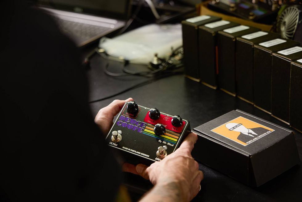 Person holding a guitar effects pedal with knobs, next to a black box and equipment.