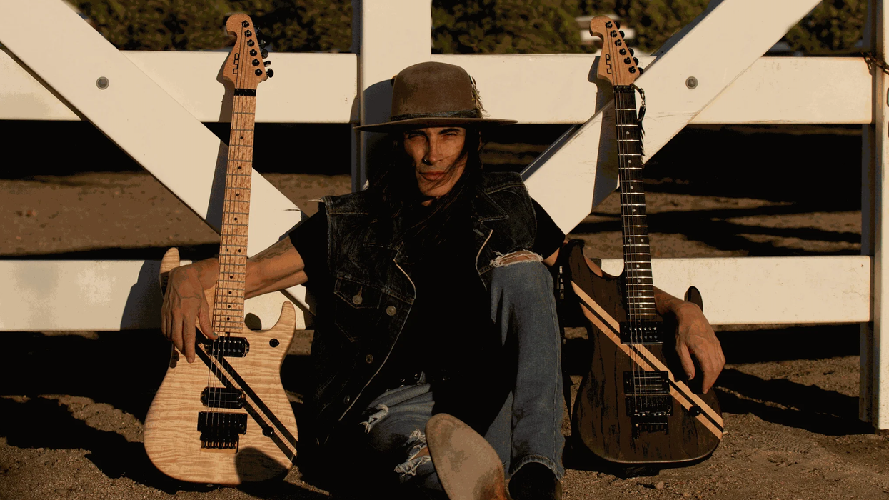 Person in denim sits by a white fence, flanked by two electric guitars, wearing a hat.