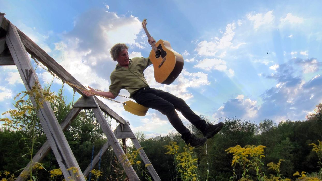 Person swinging on a playground, playing an acoustic guitar under a sunny sky.